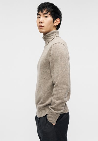 Μπεζ πουλόβερ ζιβάγκο από απαλό πλεκτό ύφασμα με ριμπ μανσέτες και τελείωμα, φορεμένο από ένα άτομο που ποζάρει πλευρικά μπροστά σε απλό φόντο.