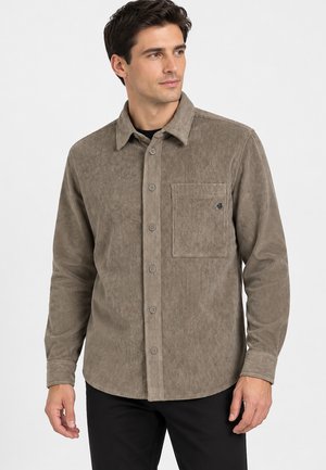 Man wearing a taupe corduroy button-up shirt with a chest pocket, looking slightly to the side, against a plain background.