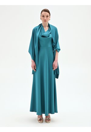 Femme portant une robe drapée bleu sarcelle avec un châle assorti, des talons ouverts dorés, posant devant un fond blanc uni.