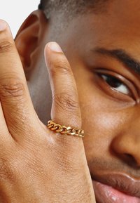 Anello in oro su un dito, con un design a catena liscio e intrecciato. Il dito presenta una pelle texturizzata e in background si intravede un primo piano di un volto.