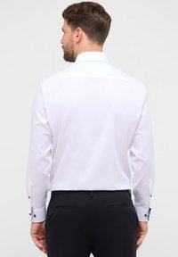 Camicia bianca con maniche lunghe, colletto classico e bottoni a contrasto sui polsini. Tessuto liscio con vestibilità sartoriale.