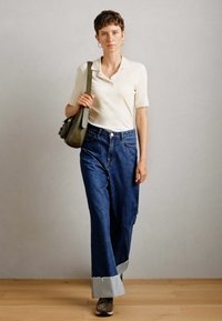 Person walking indoors wearing a cream short-sleeve top, wide cuffed blue jeans, gray sneakers, and carrying an olive green shoulder bag.