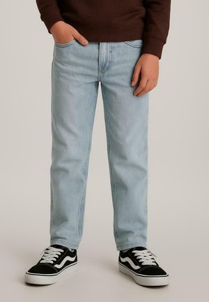 Persoon draagt lichtblauwe spijkerbroek, zwart-witte sneakers en een bruin shirt met lange mouwen, met één hand in de zak en de andere ontspannen langs het lichaam.