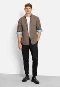 Mann trägt ein braun kariertes Hemd mit hochgekrempelten Ärmeln über einem weißen T-Shirt, schwarze Hose und schwarze Slipper und steht an einer weißen Wand.