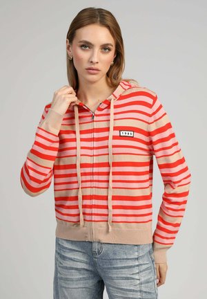 Femme portant un sweat à capuche zippé avec des rayures horizontales rouges et beiges ainsi qu'un patch "1985", associée à un jean bleu clair délavé.