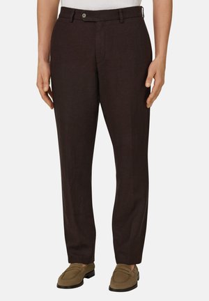 Mann trägt dunkelbraune, maßgeschneiderte Hose und olivgrüne Loafers, steht vor einem schlichten hellen Hintergrund.