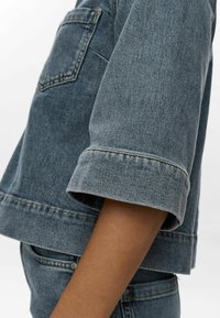 Jeans jakna v srednji modri barvi, s krojena oblika, zavihanimi rokavi in žepom na prsih z vidnimi šivi.