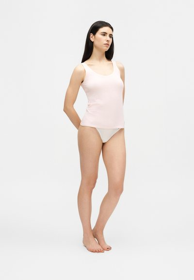 Fiatal nő hosszú sötét hajjal, világos rózsaszín ujjatlan felsőt és fehér fehérneműt visel, mezítláb áll egy egyszerű fehér háttér előtt.