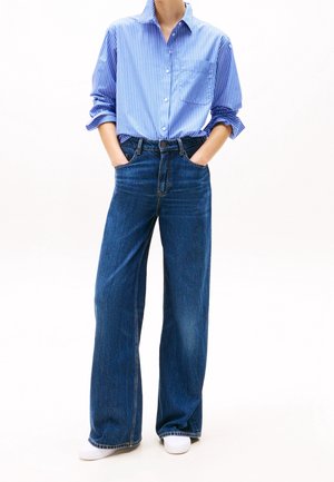 Camicia a bottoni a righe azzurro chiaro; jeans di denim blu scuro a vita alta e gamba larga; scarpe bianche; mani nelle tasche.