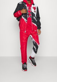 Rød og sort atletisk outfit bestående af en jakke med abstrakt print, matchende bukser og en rød hættetrøje med logo. Sorte sneakers.