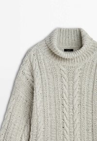 Pull en tricot épais couleur crème avec motifs torsadés et col roulé plié.