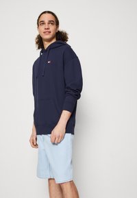 Marineblaue Kapuzenjacke aus weichem Stoff, mit einer Fronttasche und einem kleinen Logo. Getragen mit hellen Denim-Shorts.