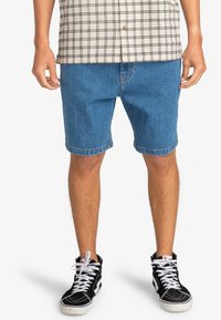 Blauwe denimshorts met een relaxte pasvorm, omgeslagen zoom, en contrasterende stiksels, gecombineerd met zwarte high-top sneakers en een geruit overhemd met knopen.