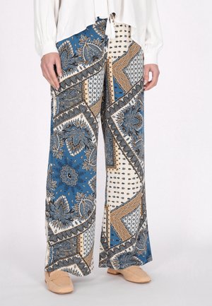 Weite Hosen mit einem blauen, weißen und goldenen Paisleymuster sowie geometrischen Mustern, aus leichtem Stoff gefertigt. Dazu beige Schuhe.