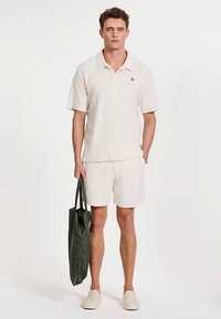 Jasnobeżowa koszulka polo, pasujące szorty, lekką ciemnozieloną torbę na ramię oraz jasnobeżowe wsuwane buty. Luźny letni strój.