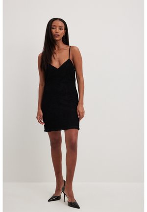 Vrouw draagt een zwarte, gestructureerde mini-jurk zonder mouwen met dunne bandjes en zwarte, puntige hoge hakken, staand tegen een effen lichte achtergrond.