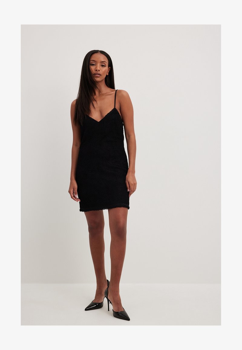 Femme portant une mini-robe noire texturée sans manches avec de fines bretelles et des escarpins noirs à bout pointu, debout devant un fond clair uni.