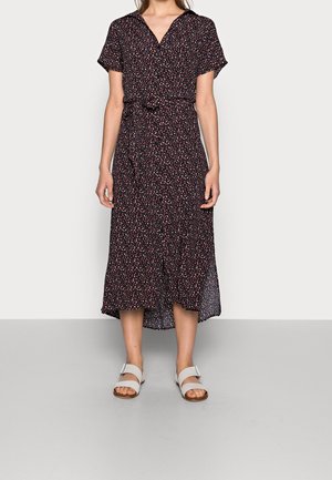 Kobieta ubrana w czarną sukienkę midi z drobnym czerwono-białym kwiatowym wzorem, krótkimi rękawami i białymi sandałami na klamrę, stojąca na szarej podłodze.