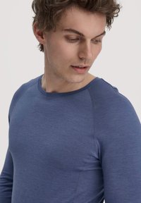 Jonge man met kort, krullend haar die een nauwsluitend blauw shirt met lange mouwen draagt en naar beneden en naar links kijkt tegen een effen lichte achtergrond.