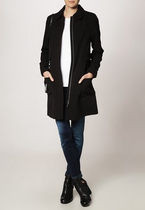 Cappotto lungo nero con chiusura a zip, tasche laterali e colletto. Indossato sopra una camicia bianca e abbinato a jeans blu scuro e stivali neri.