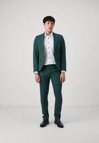 Costume vert avec une veste structurée et un pantalon ajusté. Chemise blanche avec des boutons noirs. Le modèle porte des chaussures noires. Fond uni.