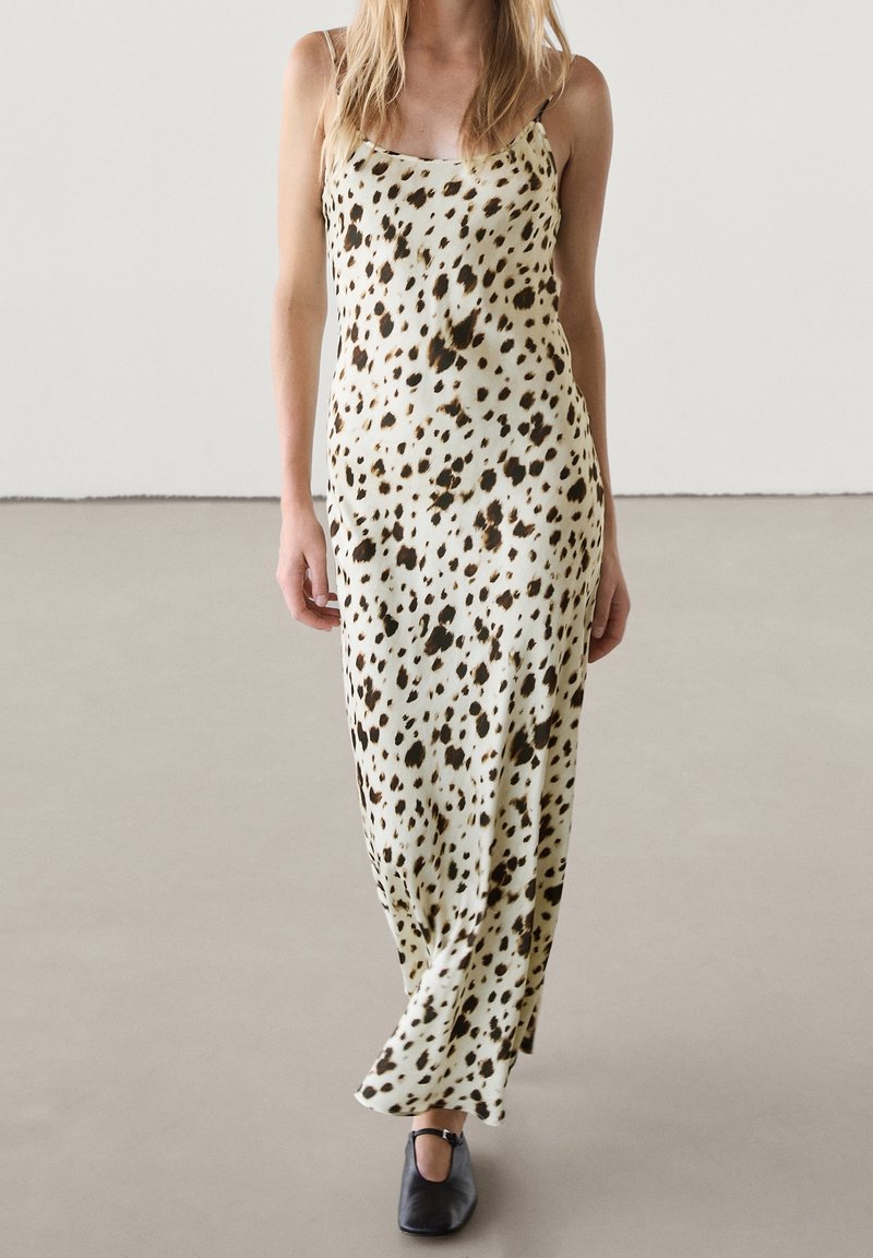 Femme portant une robe crème sans manches, longue jusqu’aux chevilles, avec des taches noires et marron, marchant sur un sol gris clair uni avec des chaussures noires.
