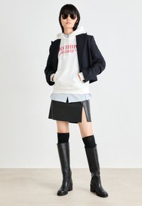 Vitt hoodie med röd logotyp, lager på lager med en ljusblå skjorta, en svart läderkjol med slits på sidan, svarta knälånga stövlar och solglasögon.