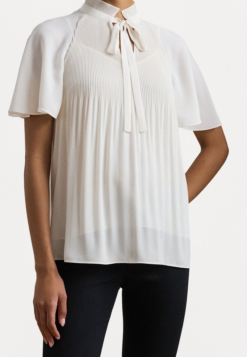 Blouse blanche avec un devant plissé, un col haut et un nœud en ruban. Manches courtes et coupe ample en tissu lisse et léger.