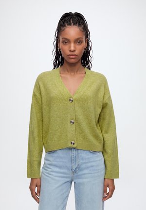 Femme aux cheveux tressés porte un cardigan vert boutonné et un jean taille haute bleu clair sur un fond blanc uni.
