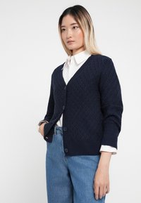 Cardigan de malha azul-marinho com textura de diamante, decote em V e cinco botões, combinado com uma camisa branca e jeans azuis.