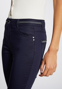 Jeans skinny azul marino con una textura suave, bolsillo con cremallera y herrajes plateados, y remaches decorativos en el bolsillo lateral.