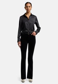 Chemise noire en satin à manches longues avec des boutons, associée à un pantalon évasé en velours noir et des chaussures blanches à imprimé serpent. Accessoires minimalistes.