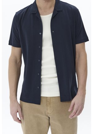 Mand iført en åben mørkeblå kortærmet skjorte over en hvid ribstrikket tanktop og lysebrune bukser, stående mod en hvid baggrund.