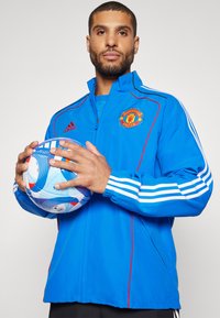 Modra športna jakna z rdečimi poudarki in belimi črtami, ki ima logo Manchester United in zadrgo spredaj. Model drži večbarvno nogometno žogo.