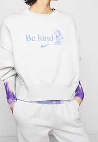 Ljusgrå sweatshirt med blå "Be Kind"-text och blommig grafik. Har rund halsringning, avslappnad passform och ribbade ärmslut, samt lila tie-dye-accenter.