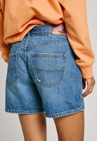 Shorts en denim bleu clair avec un effet délavé, dotés de deux poches arrières et d'une étiquette de marque en cuir à la taille. Matériau en coton, design décontracté.