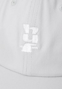 Light grey cap made of fabric, featuring a white embroidered logo design. The brim is slightly curved, with visible stitching details.