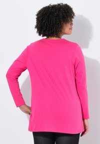 Frau mit lockigem Haar, die ein leuchtend pinkes langärmliges Shirt trägt, mit der Hand auf der Hüfte stehend und einer weißen Wand zugewandt.