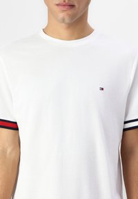 Hvid bomuld T-shirt med rund halsudskæring og korte ærmer med røde og marineblå stribede detaljer. Lille logo på brystet.