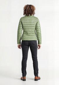 Lichtgroene pufferjack met een geborduurd ontwerp, hoge kraag en elastische manchetten, gecombineerd met donkere jeans en bruine schoenen.