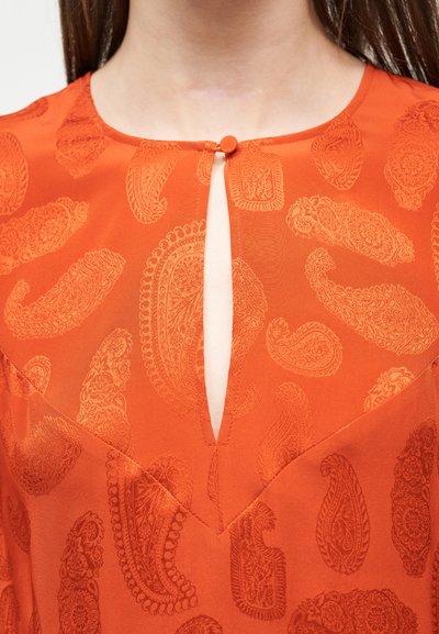 Πορτοκαλί μπλούζα με σχέδιο παϊέτες, στρογγυλή λαιμόκοψη και μικρή μπροστινή σχισμή που κλείνει με ένα κουμπί καλυμμένο με ύφασμα.