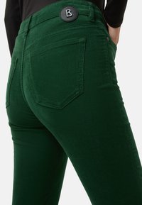 Zelené manšestrové džíny s přiléhavým střihem, s kapsami na zadní straně a černým knoflíkem s logem v pase. Texturovaná žebrovaná látka.