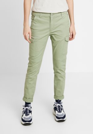 Persoon die lichtgroene cargobroek met zijzakken, opgerolde boorden, witte en marineblauwe sneakers en een wit topje draagt, staand tegen een effen achtergrond.