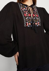 Svart blus med lös passform, med en dekorativ broderad halsringning i röd, blå och grön. Utsmyckad med en knytning och fransar.