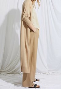 Femme portant un long cardigan beige, un pantalon large beige, un haut clair et des sandales noires à enfiler, debout devant un fond en tissu blanc drapé.