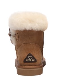 Brown suede boot with a fluffy cream shearling cuff, featuring a triangular brown logo patch at the back and a flat rubber sole.