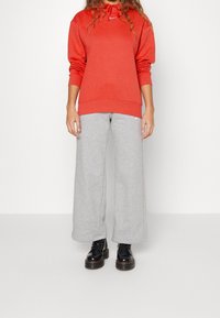 Rode sweatshirt met capuchon en lange mouwen, gecombineerd met lichtgrijze wijde sweatpants en zwarte veterschoenen. Eenvoudige, casual sportkleding.