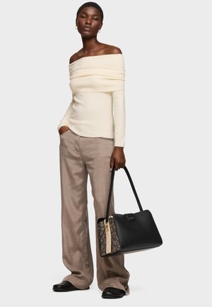 Woman wearing an off-shoulder cream top, loose beige pants, black shoes, and holding a black and patterned handbag by her side.