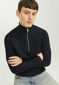 Marineblauer Strickpullover mit hohem Kragen und Viertel-Reißverschluss, mit strukturierten Kabelmustern und gerippten Bündchen. Kombiniert mit hellen Jeans.