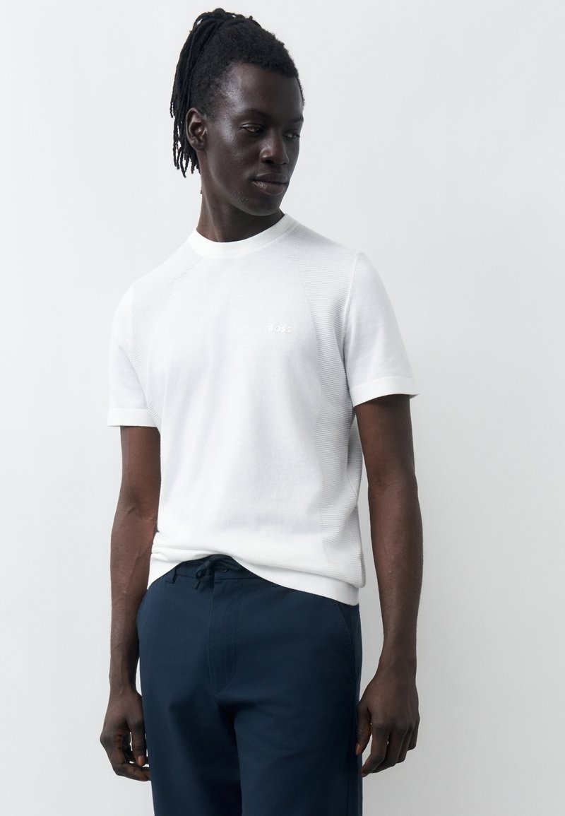 Άνδρας με σκούρο δέρμα και dreadlocks φοράει λευκό πλεκτό πουκάμισο με κοντά μανίκια και ναυτικό μπλε παντελόνι, στέκεται μπροστά σε απλό φωτεινό φόντο.